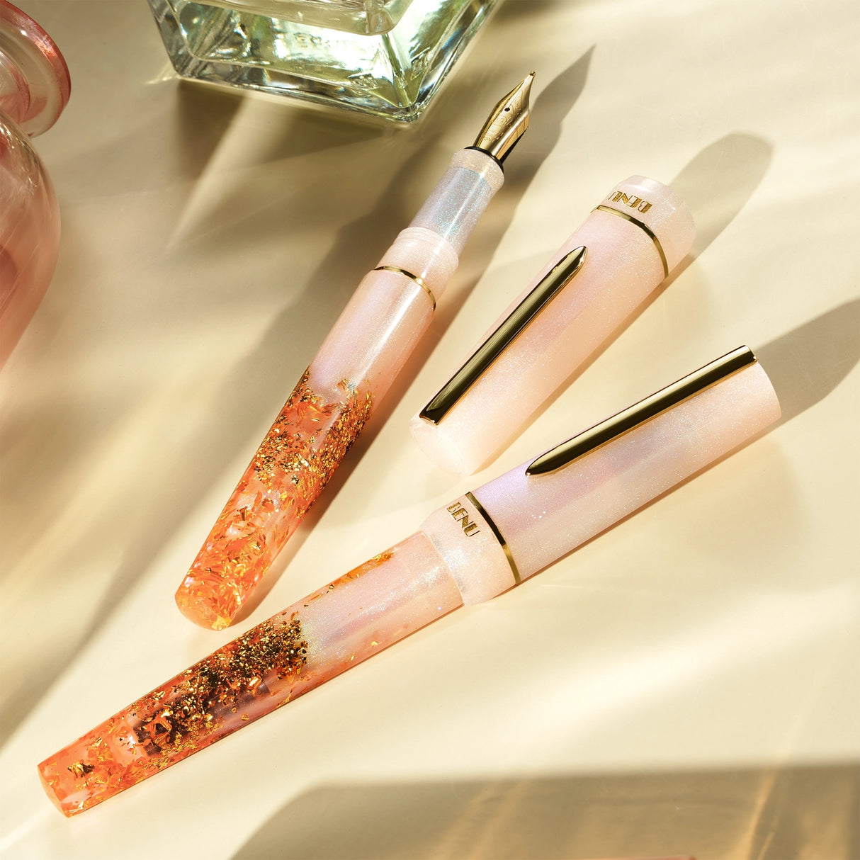 BENU presents the newest Haute Collection — a line of elegant, fashion-inspired pens that blend classic charm with bold, modern flair. The designs include both subtle and vibrant styles, showcasing BENU’s signature glitter and color artistry. Each pen features a faceted body with alternating wide and slender facets, complemented by either gold-plated or chrome-plated trims for a sleek, timeless look.
A mix of golden flakes dances through a soft, pearly pink base, complemented by gleaming gold trim. Tender