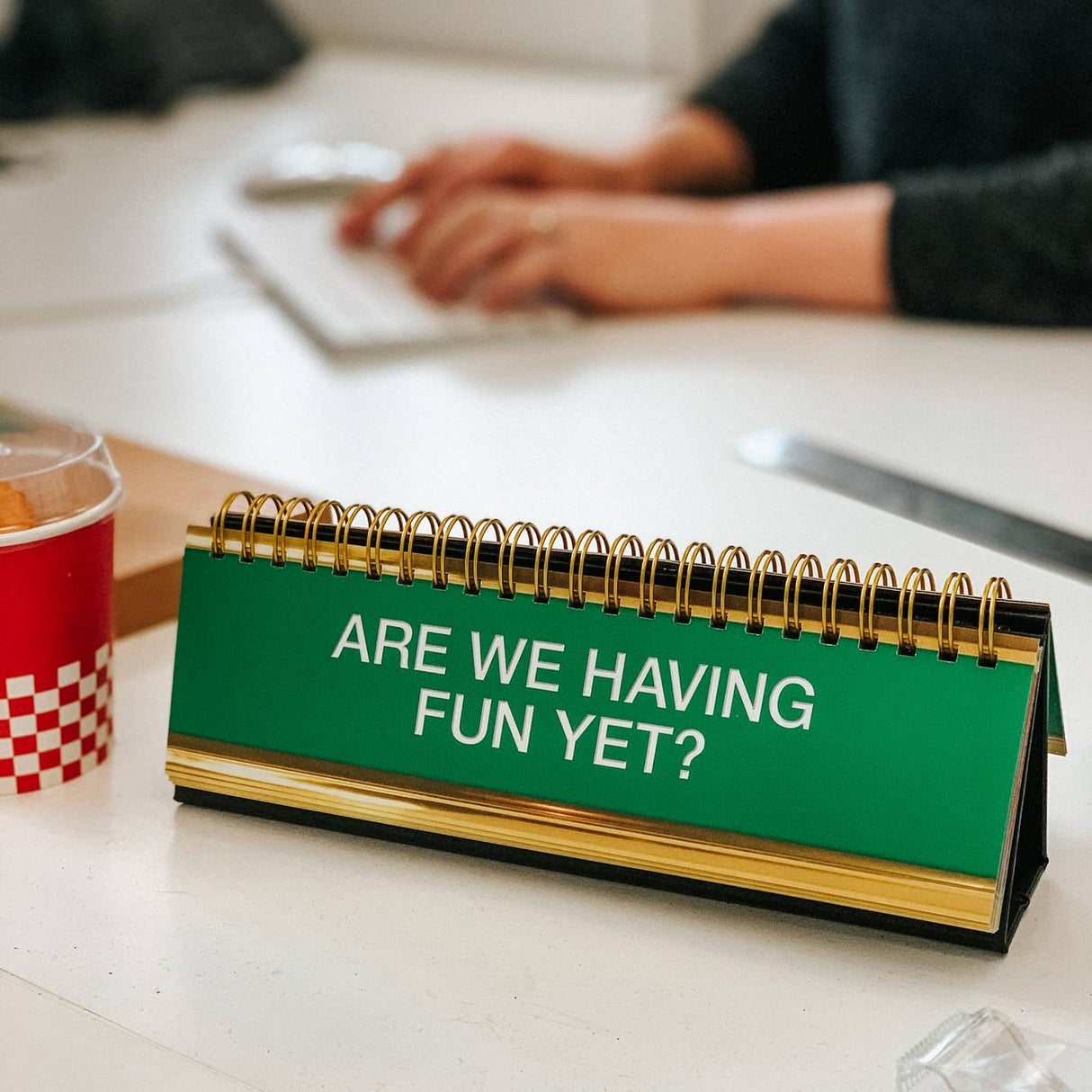 Desk calendar with 'Are we having fun yet?' text on a desk with a blurred background of a person working.