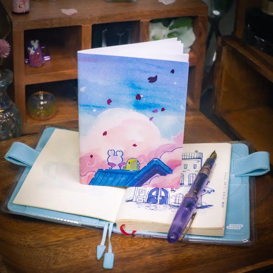 Esterbrook x Sakuradragon - Sakura Street Morning Skies A6 Notebook
Mornings are quiet around here.
Did you notice the pink foil cherry blossoms falling from the sky?
Dimensions: A6 (10.5 × 14.8 cm)
Paper: Double-sided offset paper
Page Ruling: Blank
Pages: 64
Message from sakuradragon: Not all paper and ink are made equal but this paper does a great job with both fountain pens and dip pens. There is minimal ghosting but bleeding does occur with heavy ink usage.