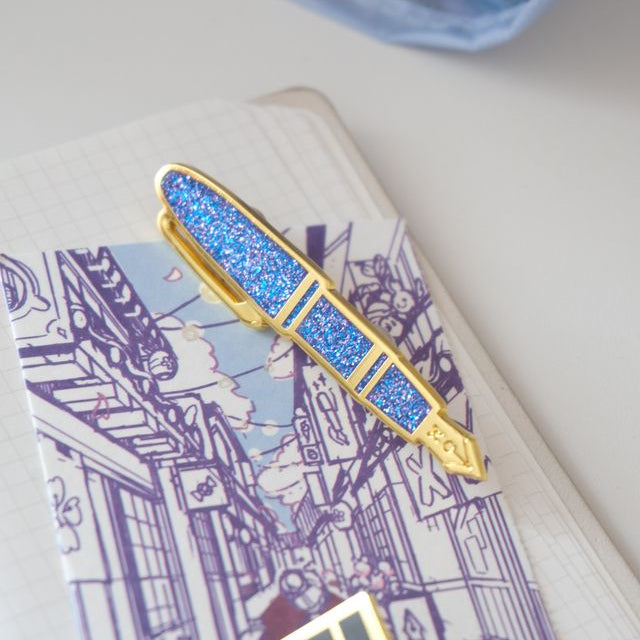 Carry the Sakura Street Estie beyond the page with this oversized hard enamel lapel pin. The 7.5 cm gold- plated design recreates the fountain pen with a sparkle effect inspired by its translucent purple material. It brings one of the collaboration's central designs to jackets, bags, and pin collections.