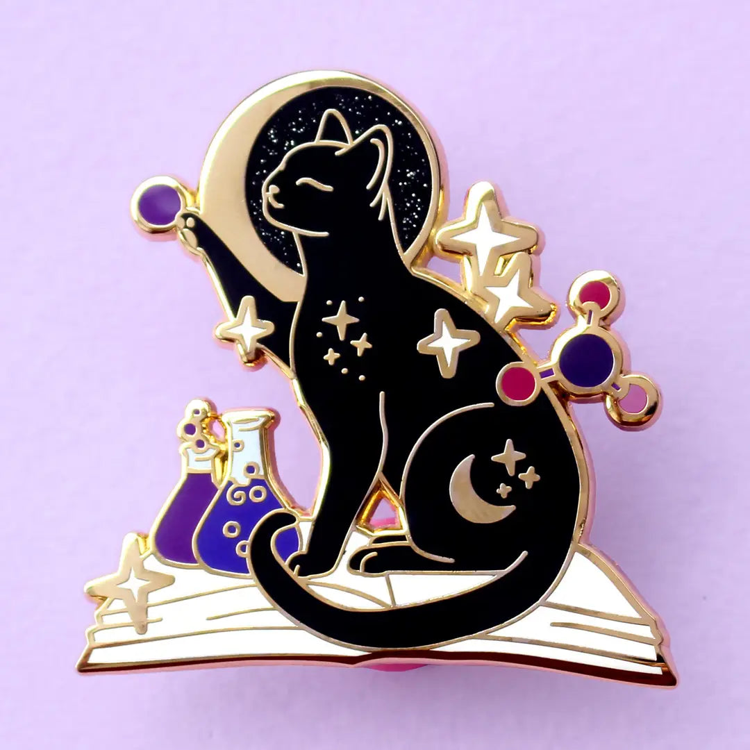 If you’re passionate about science and partial to cats, this awesome pin is perfect for you! It features a black cat sitting on a book and playing with atom parts. It also features a glittery moon and two beakers filled with purple liquid.
The pin is 42mm (1.65”) tall, and is made from gunmetal hard enamel, with glitter and UV printed details. It's secured with 2 pink clutches to keep the pin in place.
