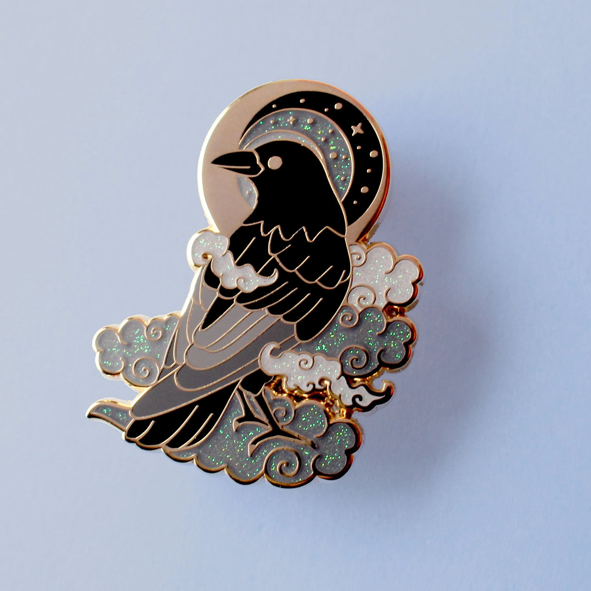 This enamel pin features a black crow surrounded by grey glittery clouds, and a starry moon with grey backgrounds. The pin is presented on a Glitter Punk backing card and is secured with 2 pin backs.
This pin is part of the Corvid Collection, an 8-part series of bird pins.
Measurements: 39mm