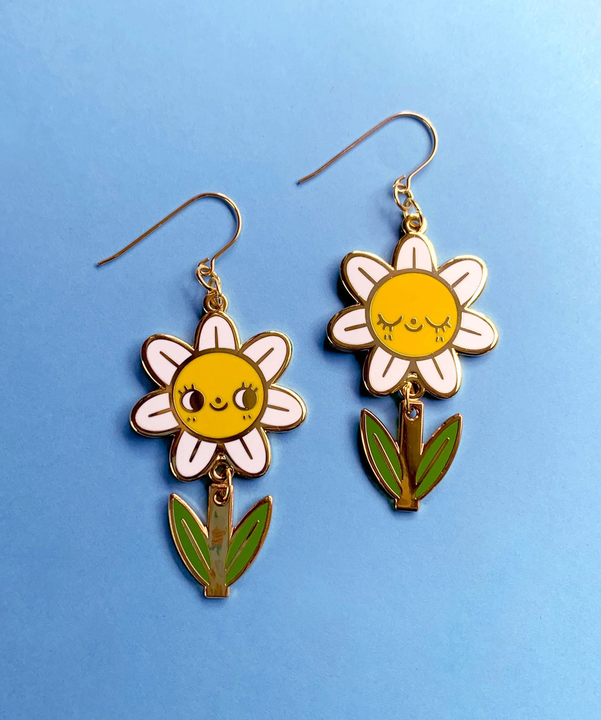 A pair of mis-matched happy daisy themed earrings (one one with happy open eyes, and one with happy closed) with separate dangling stems. Made from hard enamel with shiny gold metal finish on high quality Hypo-Allergenic, gold plated, brass based hooks.
Each earring measures approximately 5cm tall x 3cm wide. Earrings come in a Hannakin embossed gift box.