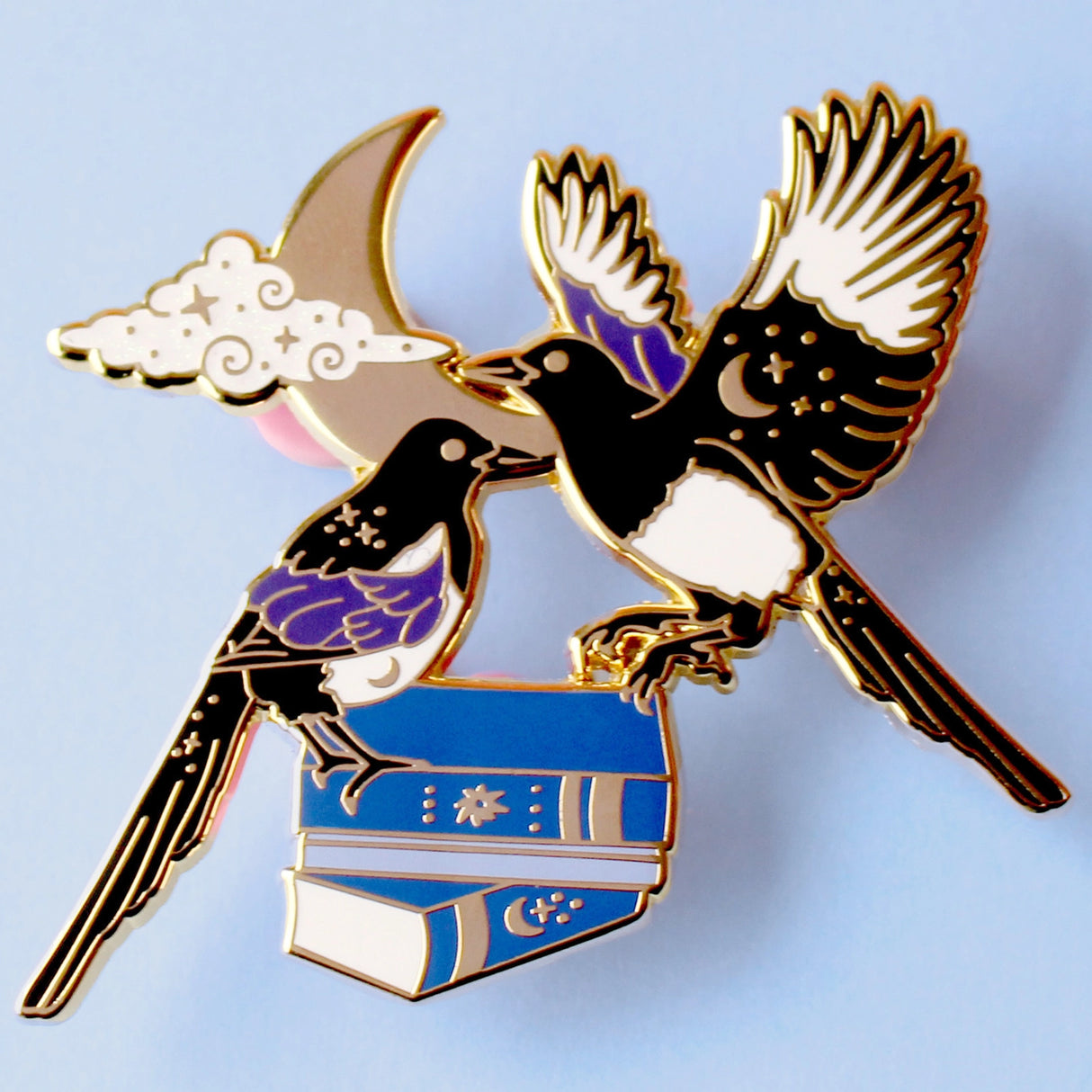 This enamel pin features a pair of magpies (2 for luck!) on top of a set of blue books, in front of a moon and glittery cloud. The pin is presented on a Glitter Punk backing card and is secured with 2 pin backs.
This pin is part of the Corvid Collection, an 8-part series of bird pins.
Measurements: 55mm
