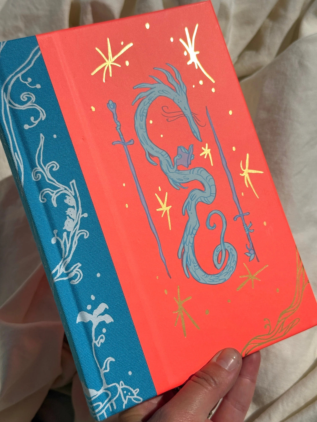 This is a blank 200 page notebook/sketchbook with thin ivory Munken paper perfect for light sketching or writing. Part of a fantasy collection called Woodfolk.
The cover is fluorescent pink/orange and has a smooth matte finish with gold foiling on the front and back. Plus it has a blue fabric spine with white embossing, adding to the charm of the book. Best for pencils, light pens, and generally light mediums, as thick markers or water can bleed. Not recommended for gouache or heavy marker use.