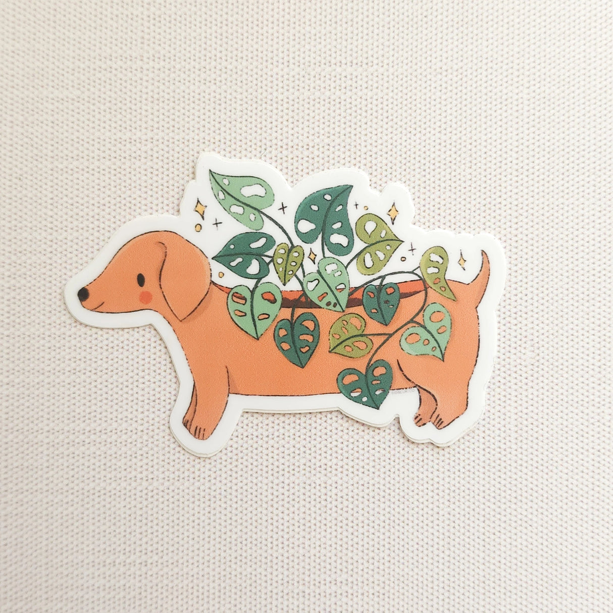 A charming dachshund planter rests among leafy greenery, a thriving cheese plant growing from its back like a small piece of playful décor. This vinyl sticker captures that cozy, plant-filled still life, adding a touch of warmth and whimsy to your everyday essentials.
Made from durable, waterproof vinyl, this sticker is designed to hold up wherever you place it—whether that’s a water bottle, laptop, or journal you reach for daily.