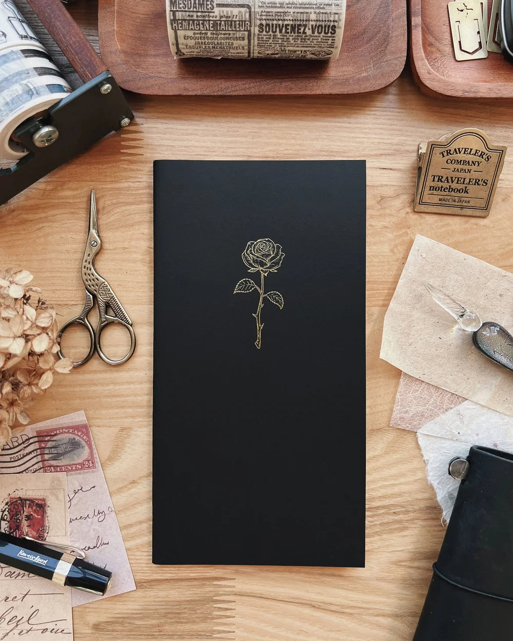 Black notebook with a rose design on a wooden surface with stationery items.