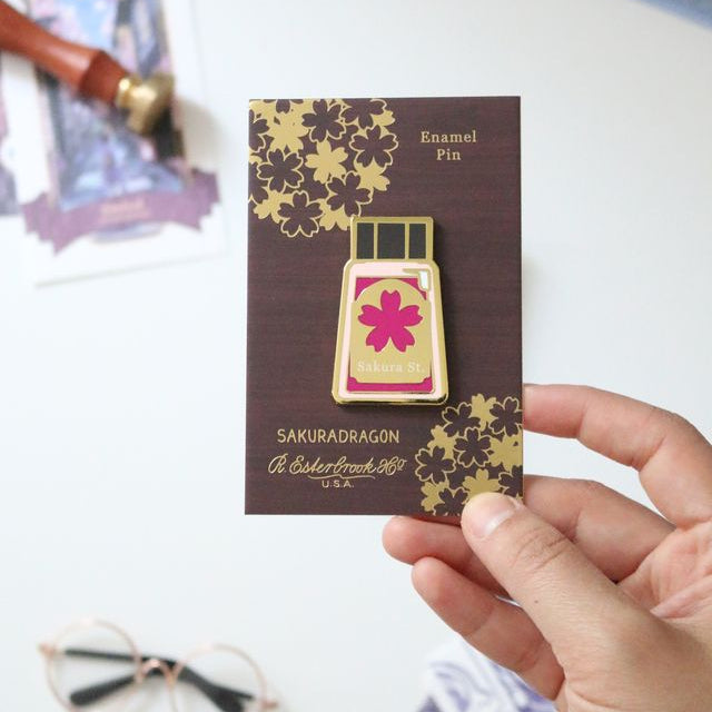 The Sakura Street Ink Lapel Pin turns one of the collection's coordinating ink bottles into a collectible hard enamel design. Measuring 4.5 cm and finished with gold-plated details, the pin carries the colors and imagination of Cindy Duong's Sakura Street artwork onto a jacket, bag, or display board.
