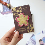 Inspired by the blossoms found throughout Cindy Duong's Sakura Street illustrations, this oversized hard enamel pin features a sakura flower outlined in gold-plated metal. The 5 cm design includes Esterbrook and Sakuradragon branding at its center, creating a collectible emblem of the collaboration.