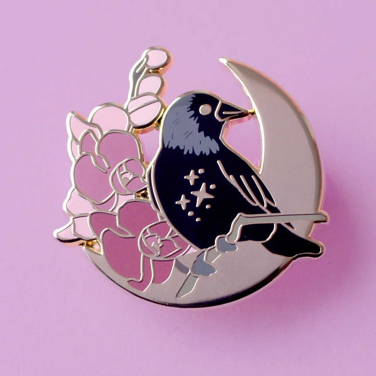 This enamel pin features a Jackdaw perching on a branch of pink orchid flowers, in front of a large gold moon. The pin is presented on a Glitter Punk backing card and is secured with 2 pin backs.

This pin is part of the Corvid Collection, an 8-part series of bird pins. 

Measurements: 35mm