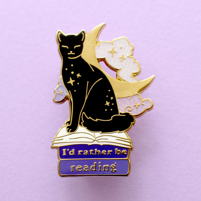 We’ve all sometimes wished we were at home with a hood book, rather than having to deal with the real world! This super cute pin features a black cat sitting on pink &amp; purple books with the slogan ‘I’d rather be reading’. Behind the cat is a large crescent moon and a pair of glittery pastel clouds.

Measurements: 45mm (1.77”)