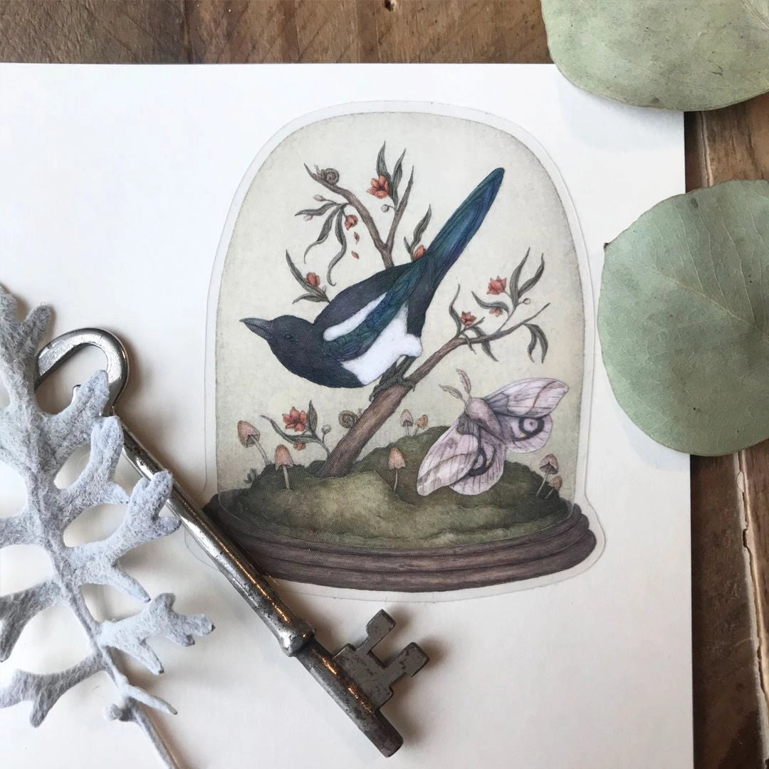 Vinyl semi-transparent sticker of One for Sorrow, an illustration of a magpie and moth under a cloche jar, by Jessica Roux.

The glass of the jar is transparent, but the contents and base of the jar are opaque. Sticker measures 2.6" x 3.1" and is glossy.