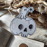 A little ghostie with a flower, perched atop a spooky skull, on a die-cut vinyl sticker. From original ink artwork by Jon Turner.