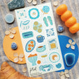 🩴🍊 @papergrapeprints is serving up a sun-soaked spread of seaside favorites, and we’ll take one of everything! Bright oranges, sardines, pasta, pretty ceramics, hydrangeas, and that perfect blue crab come together in a dreamy mix of coastal color and vacation-day charm. Add flip-flops, something cold to sip, and absolutely zero plans. Art by: @papergrapeprints

Top Coating: None
Adhesive Layer: Paper
Backing Layer: Clear PET
6.69" x 3.54".
Weight - 0.03lbs
1 sheet included
