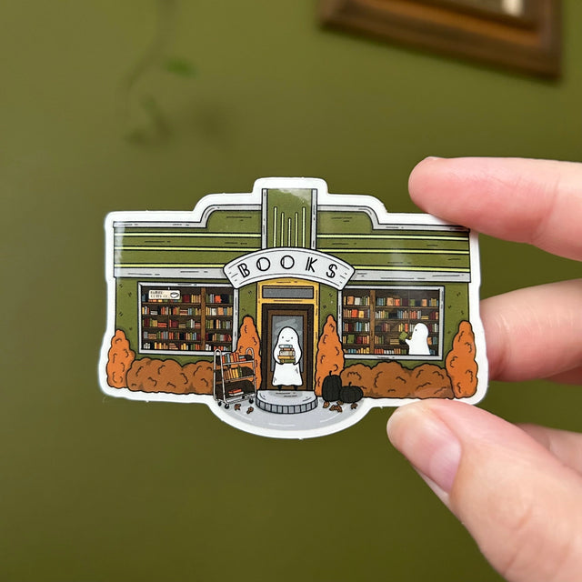 Glossy sticker of a little art deco bookshop where a ghost with a stack of books stands in the doorway.

3in wide
Waterproof
Glossy finish