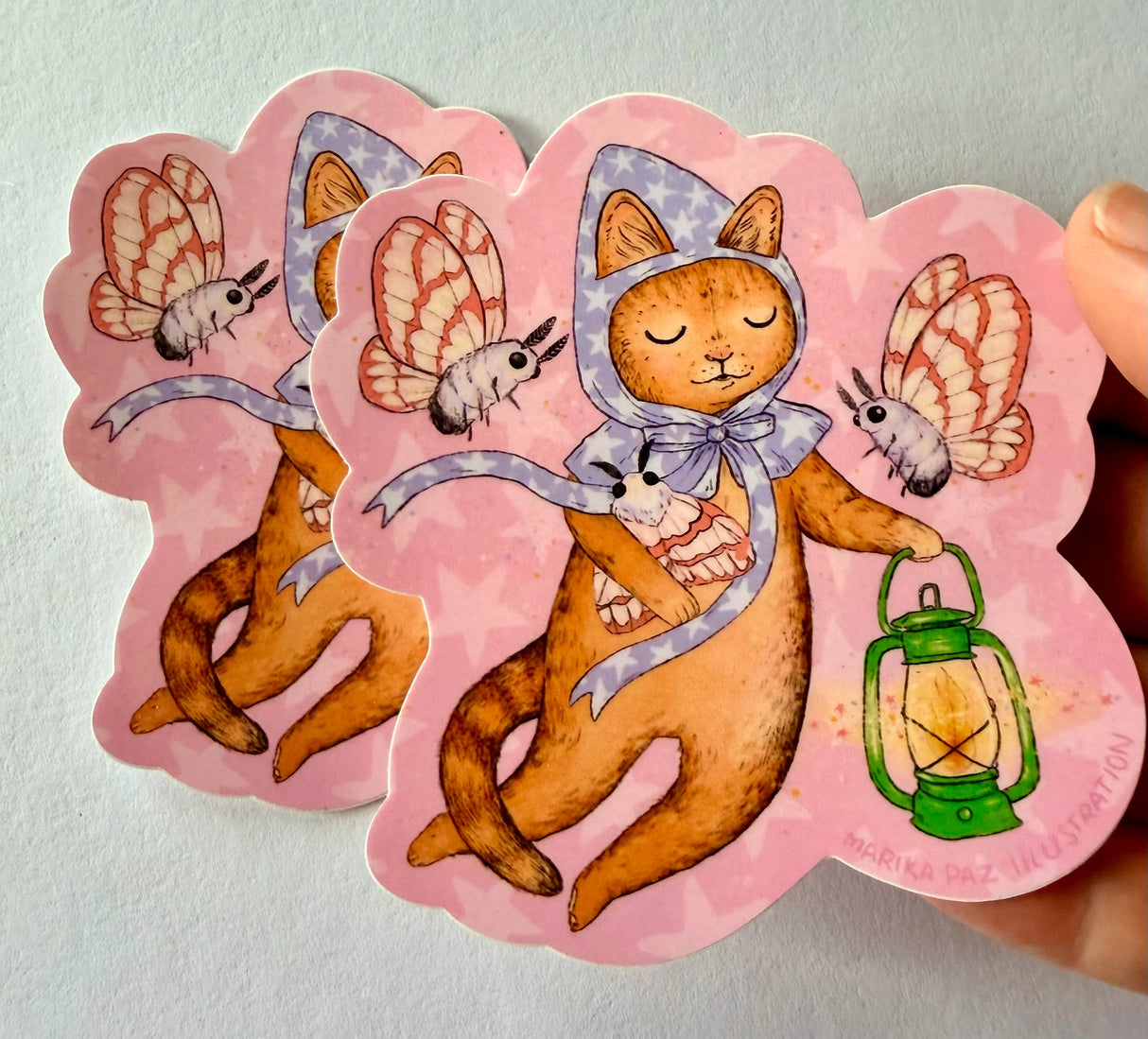 A cat and his lantern attract moths as they float amongst the stars.

This vinyl matte sticker is weather and waterproof-stick it on a notebook, bike, or car- it's going to stick around! Heck, throw it on a water bottle and through the dishwasher. This sticker can go anywhere. Sticker measures 3.5" x 4.5" Printed in USA.
