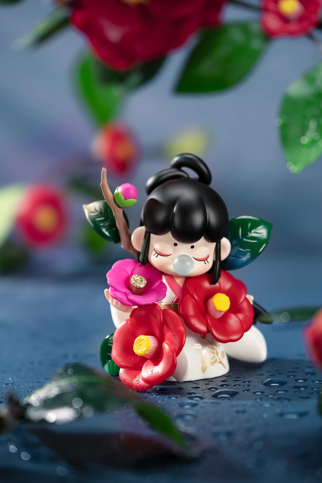 Nanci Flower Stories Figure Dolls Blind Box Small figurine of a girl with black hair and floral dress on a blue surface with flowers and leaves.