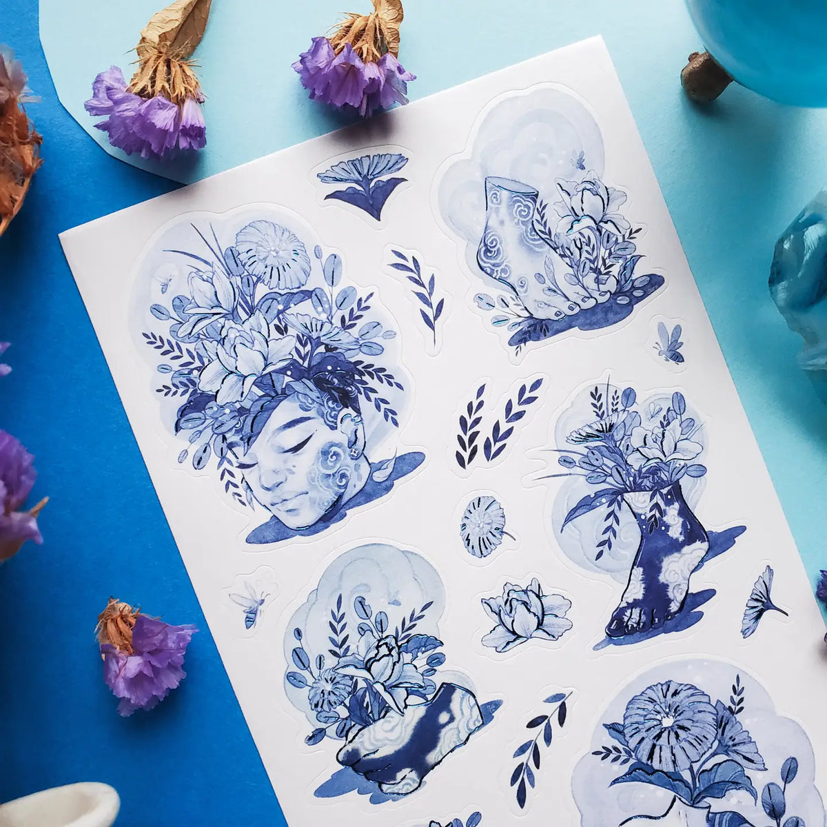 💐🌀 Step into @so.meurdok’s surreal garden, where faces, hands, feet, and figures blossom into living bouquets. Lush florals and striking blue details blur the line between body and bouquet, while foil accents trace every elegant curve like enchanted brushstrokes. A tiny art gallery, now in sticker form. Art by: @so.meurdok

Top Coating: Blue foil
Adhesive Layer: Paper
Backing Layer: Paper
6.69" x 3.54".
Weight - 0.03lbs
1 sheet included