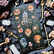 In @iratxe.yc’s shadowed garden, nothing is quite as it first appears. Jeweled flowers gaze back from curling stems, delicate moths wear watchful eyes, and a silver-haired guardian stands adorned in blossoms, gemstones, and armor. Rich with surreal beauty and dark botanical magic, this striking sheet reveals a forest where every petal may be hiding a secret.

Art by: @iratxe.yc

One sticker sheet is included.
Top Coating: None
Adhesive Layer: Paper
Backing Layer: Clear PET
Size:6.69"x3.54"
Weight-0.03oz