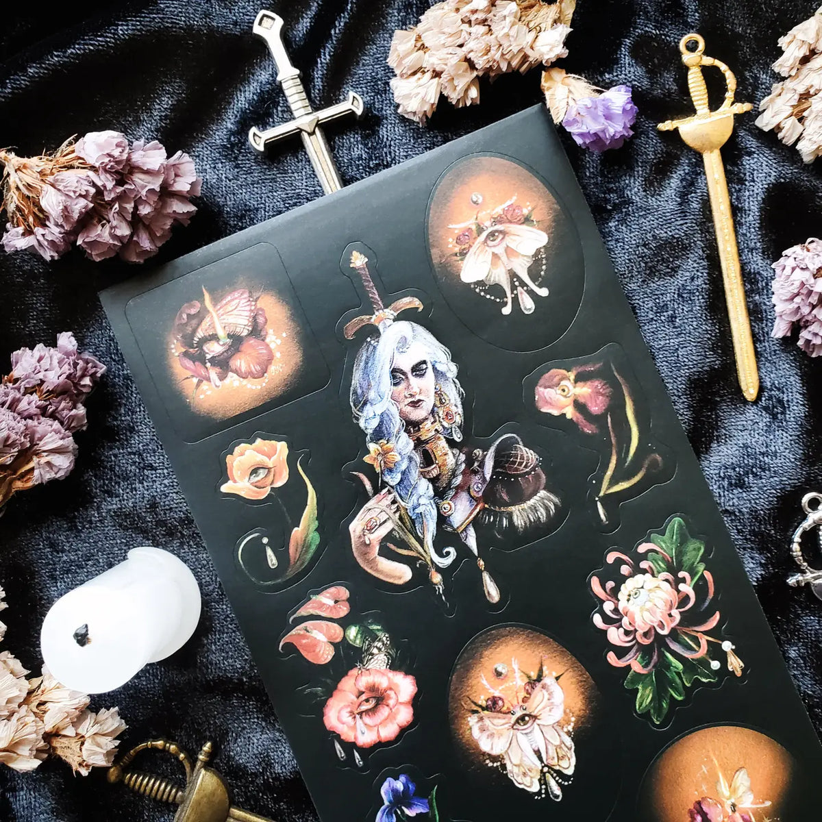 In @iratxe.yc’s shadowed garden, nothing is quite as it first appears. Jeweled flowers gaze back from curling stems, delicate moths wear watchful eyes, and a silver-haired guardian stands adorned in blossoms, gemstones, and armor. Rich with surreal beauty and dark botanical magic, this striking sheet reveals a forest where every petal may be hiding a secret.

Art by: @iratxe.yc

One sticker sheet is included.
Top Coating: None
Adhesive Layer: Paper
Backing Layer: Clear PET
Size:6.69"x3.54"
Weight-0.03oz